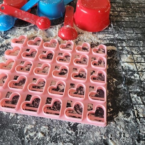 May include: A pink heart-shaped cookie cutter with 36 individual heart-shaped molds. The cutter is on a black surface covered in white flour.