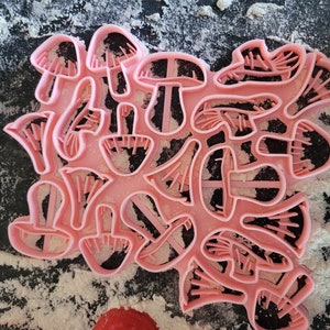 May include: A set of pink plastic cookie cutters shaped like mushrooms. The cutters are arranged in a cluster on a surface covered in white powder.