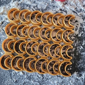 May include: A gold-colored plastic cookie cutter with a crescent moon shape. The cutter is made up of multiple crescent moons arranged in a grid pattern.