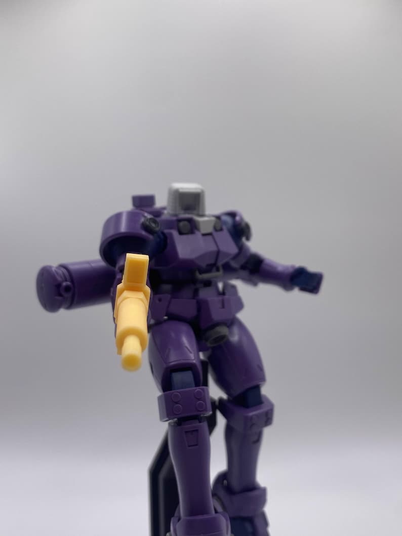 May include: A purple and gray robot toy holding a yellow gun. The robot has a detailed design with multiple joints and is posed in a dynamic action pose.