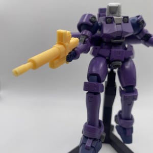 May include: A purple robot action figure holding a yellow plastic gun. The robot is standing on a black stand.