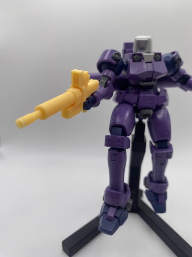 May include: A purple robot action figure holding a yellow plastic gun. The robot is standing on a black stand.