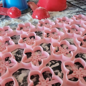 May include: A set of pink plastic cookie cutters shaped like cherry blossoms. The cutters are arranged in a grid pattern on a surface covered in white flour.