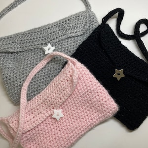May include: Three handmade crochet purses in gray, black, and pink. Each purse features a flap closure and a long strap. A white star-shaped button secures the flap on each bag. The purses are made of a thick yarn.