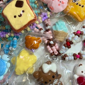 May include: A collection of assorted plastic charms and beaded accessories in clear plastic bags. The charms include a smiling toast slice, a pink creature, a yellow star, a brown bear with a white bow, and a blue ice cream cone.