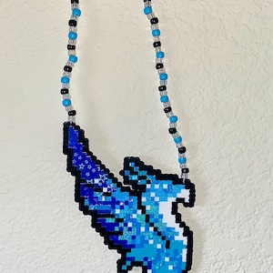 May include: A beaded necklace featuring a pixelated blue and white bird pendant. The pendant has a black outline and is made of small, square beads. The necklace chain alternates black, blue, and clear beads.