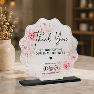 May include: A scalloped acrylic sign featuring a floral design with pink roses and the text Thank You FOR SUPPORTING OUR SMALL BUSINESS Follow us on! with social media icons and a website placeholder, mounted on a rectangular black base.