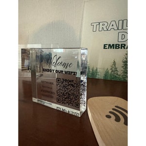 May include: A clear acrylic block with the words "Welcome Enjoy Our Wifi!" printed on the front. The block displays the network name, password, and a QR code. A wooden wifi symbol is on the right.