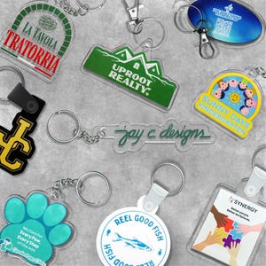 Peut inclure: Une collection de porte-clés en acrylique personnalisés présentant divers designs, notamment La Tavola Trattoria, Uproot Realty, Sunny Days Kindergarten, Jay C. Designs, Gentle Paws Clinic, Reel Good Fish et Synergy avec des logos et textes colorés.