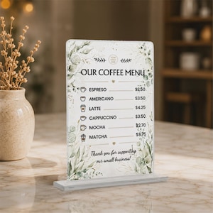 May include: A rectangular acrylic sign featuring a coffee menu with green botanical illustrations. The text reads OUR COFFEE MENU, listing espresso, americano, latte, cappuccino, mocha, and matcha with prices, ending with Thank you for supporting our small business.