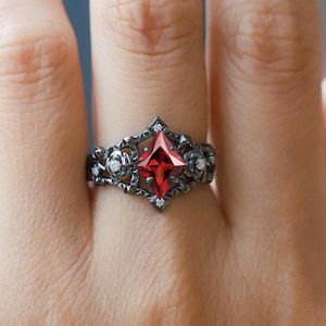 Puede incluir: Un anillo negro con una gema roja de corte cuadrado en el centro. El anillo tiene un diseño ornamentado con detalles florales y pequeñas piedras blancas. El anillo está en un dedo.