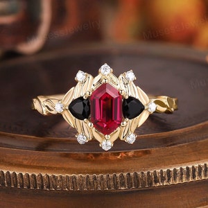 May include: A gold ring with a red gemstone and black gemstones set in a diamond-shaped halo. The ring is surrounded by smaller diamonds.