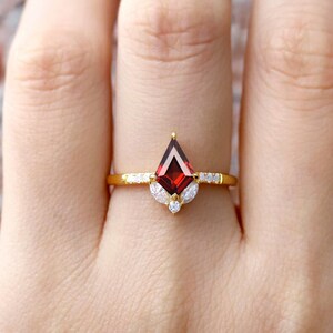 Puede incluir: Un anillo de oro con una piedra preciosa roja y pequeñas piedras blancas. El anillo se lleva en un dedo.