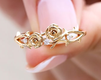 May include: A gold ring featuring two sculpted rose blossoms and thorny vine details along the band. Three small, clear, round-cut stones are set between the roses and along the vine, adding sparkle to the polished yellow gold metal finish.