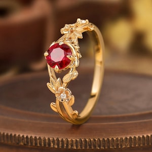 May include: A gold ring featuring a vibrant red gemstone at its center. The ring is adorned with delicate floral and leaf designs, with small accent diamonds. The ring's band and setting are a polished gold color, creating a luxurious appearance.