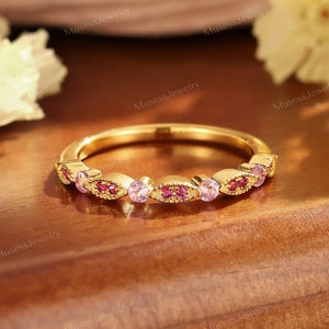 May include: A delicate gold band ring with a row of pink and red gemstones. The ring is set on a wooden surface.