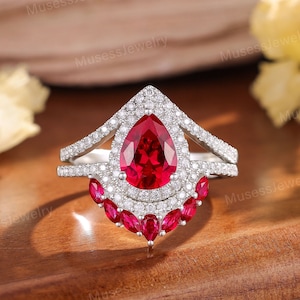 May include: A pear-shaped ruby engagement ring with a halo of smaller diamonds. The ring has a split shank with a row of smaller rubies on each side.