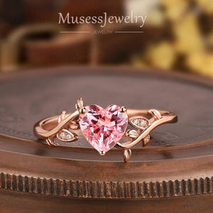 May include: Rose gold ring with a heart-shaped pink gemstone. The ring features a detailed vine design with small, sparkling accent stones. The ring is displayed on a wooden surface.