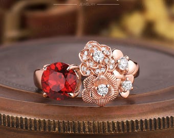 May include: A rose gold ring with a large red gemstone and smaller white gemstones set in a floral design. The ring is on a wooden surface.