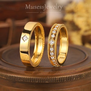 Puede incluir: Dos anillos de oro con bordes de cuentas. El anillo izquierdo presenta una piedra de corte princesa, mientras que el derecho muestra una hilera de piedras redondas engastadas en una banda de filigrana. El texto MusessJewelry es visible.