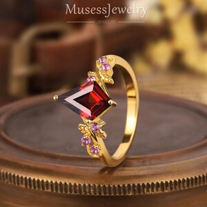 May include: A gold ring with a red square-shaped gemstone surrounded by smaller pink gemstones. The ring has a delicate floral design.