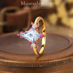 May include: A gold ring with a light blue square-shaped gemstone surrounded by smaller pink and white gemstones. The ring is set on a brown surface.