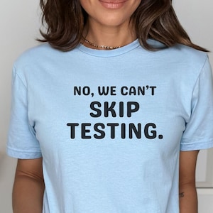 May include: Light blue t-shirt with the text "NO, WE CAN'T SKIP TESTING." in black. The shirt is a light blue color and has short sleeves. The words "Light Blue" are printed at the bottom of the image.