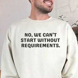 May include: A light beige sweatshirt with the text "NO, WE CAN'T START WITHOUT REQUIREMENTS." printed in black.