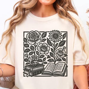 May include: Cream-colored t-shirt featuring a black and white floral design within a square frame. The design includes roses, daisies, and other flowers, along with open and closed books. The shirt is a casual, comfortable style.