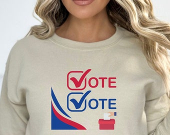 May include: A sand-colored sweatshirt with a graphic of two checkboxes with the word "VOTE" next to each. The graphic also includes a red, white, and blue ribbon and a red ballot box.