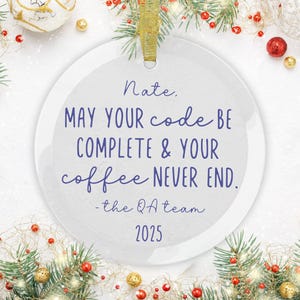 May include: A round, clear glass ornament with a gold ribbon for hanging. The ornament has the text "Nate, MAY YOUR code BE COMPLETE & YOUR coffee NEVER END. -the QA team 2025" in blue script.