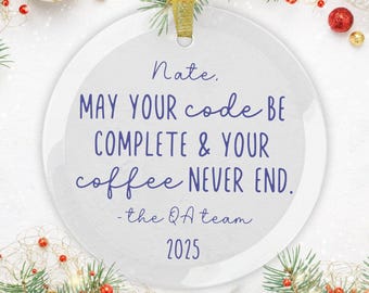 May include: A round, clear glass ornament with a gold ribbon for hanging. The ornament has the text "Nate, MAY YOUR code BE COMPLETE & YOUR coffee NEVER END. -the QA team 2025" in blue script.