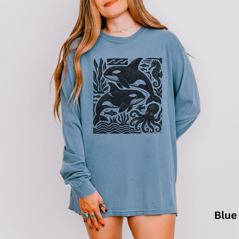 Op de afbeelding: Blauw shirt met lange mouwen met een zwart-wit afbeelding van twee orka's, vissen, een octopus en zeewier. Het ontwerp staat in een vierkant kader. Het woord "Blue" staat rechtsonder afgedrukt.