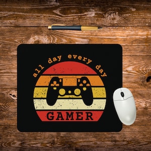 May include: Black mousepad with a retro design featuring a game controller silhouette inside a sunset-colored circle. The text "all day every day" arcs around the top, and "GAMER" is at the bottom. A white computer mouse is to the right.