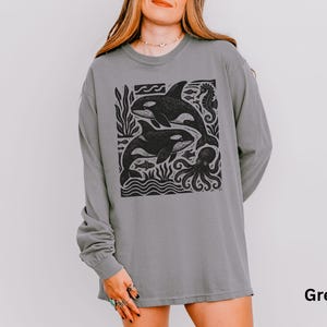 Op de afbeelding: Een grijze longsleeve met een zwarte grafische print van twee orka's, een octopus en andere zeedieren. De shirt heeft een ronde hals en lange mouwen. Het woord "Grey" staat rechtsonder gedrukt.