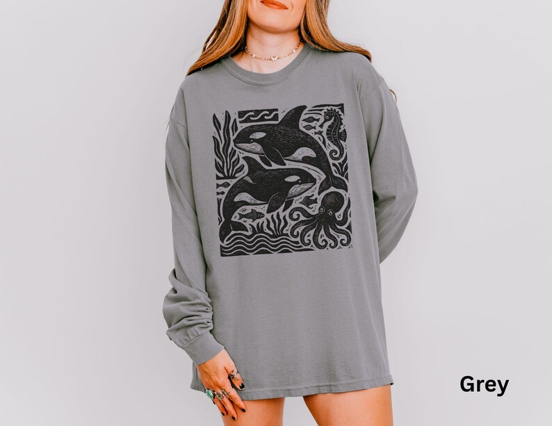 Op de afbeelding: Een grijze longsleeve met een zwarte grafische print van twee orka's, een octopus en andere zeedieren. De shirt heeft een ronde hals en lange mouwen. Het woord "Grey" staat rechtsonder gedrukt.