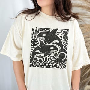 Op de afbeelding: Ivoor kleurig T-shirt met een zwart-witte afbeelding van orka's, een zeepaardje en een octopus. Het ontwerp bevat gestileerde golven en zeewier. Het shirt heeft een relaxte pasvorm.