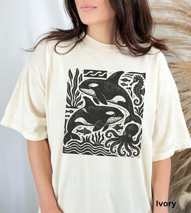 Op de afbeelding: Ivoor kleurig T-shirt met een zwart-witte afbeelding van orka's, een zeepaardje en een octopus. Het ontwerp bevat gestileerde golven en zeewier. Het shirt heeft een relaxte pasvorm.