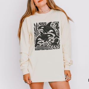 Op de afbeelding: Ivoor lange mouwen shirt met een zwart-witte afbeelding van twee orka's, zeewier en een octopus. Het shirt heeft een ronde hals en lange mouwen. Het woord "Ivory" staat rechtsonder afgedrukt.