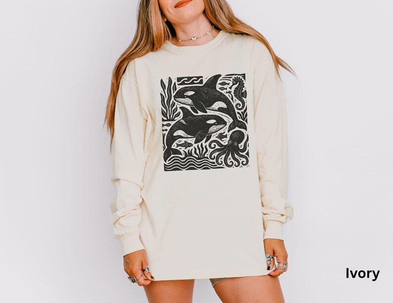 Op de afbeelding: Ivoor lange mouwen shirt met een zwart-witte afbeelding van twee orka's, zeewier en een octopus. Het shirt heeft een ronde hals en lange mouwen. Het woord "Ivory" staat rechtsonder afgedrukt.