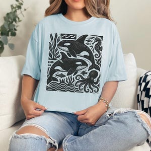 Op de afbeelding: Lichtblauw T-shirt met een zwart-witte print van orka's, een zeepaardje en een octopus. Het ontwerp bevat gestileerde golven en zeewier. Het T-shirt heeft een losse pasvorm.