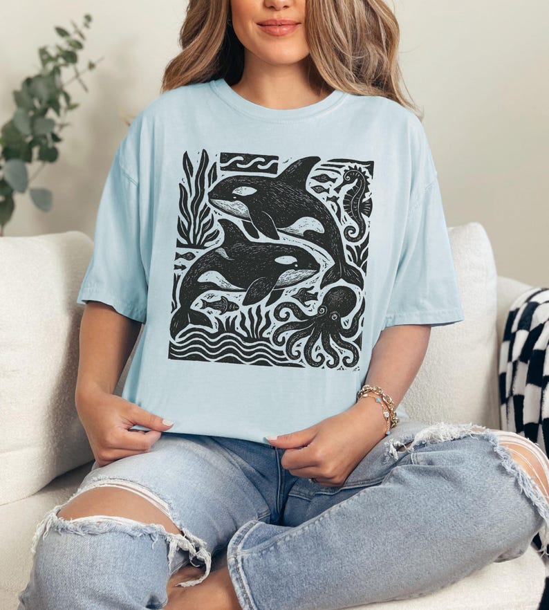 Op de afbeelding: Lichtblauw T-shirt met een zwart-witte print van orka's, een zeepaardje en een octopus. Het ontwerp bevat gestileerde golven en zeewier. Het T-shirt heeft een losse pasvorm.