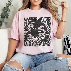 Op de afbeelding: Lichtroze T-shirt met een zwart-witte afbeelding van orka's, een zeepaardje, een octopus en ander zeeleven. De tekst "Blossom" staat onderaan het shirt.