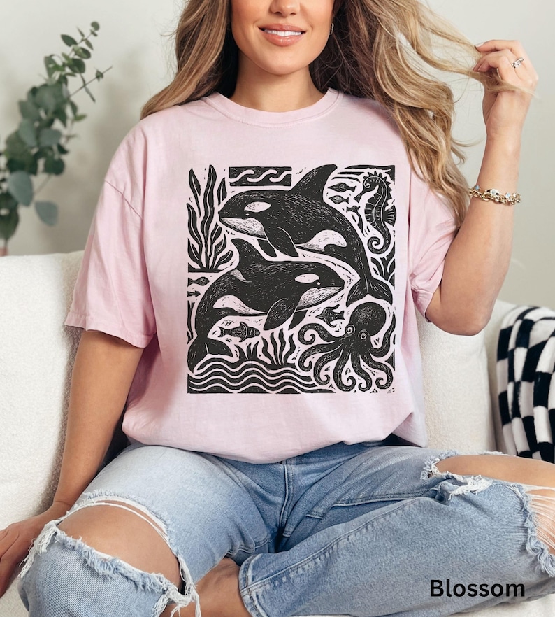 Op de afbeelding: Lichtroze T-shirt met een zwart-witte afbeelding van orka's, een zeepaardje, een octopus en ander zeeleven. De tekst "Blossom" staat onderaan het shirt.