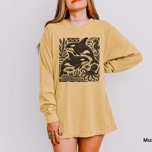 Op de afbeelding: Mosterdkleurig shirt met lange mouwen met een zwarte afbeelding van twee orka's, een zeepaardje en een octopus. De orka's zwemmen in de oceaan. Het shirt is een comfortabele, casual stijl.