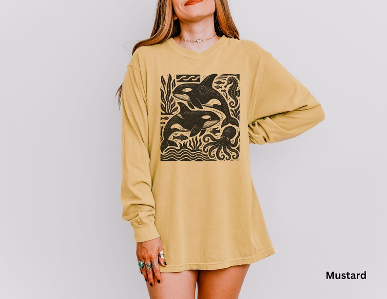 Op de afbeelding: Mosterdkleurig shirt met lange mouwen met een zwarte afbeelding van twee orka's, een zeepaardje en een octopus. De orka's zwemmen in de oceaan. Het shirt is een comfortabele, casual stijl.