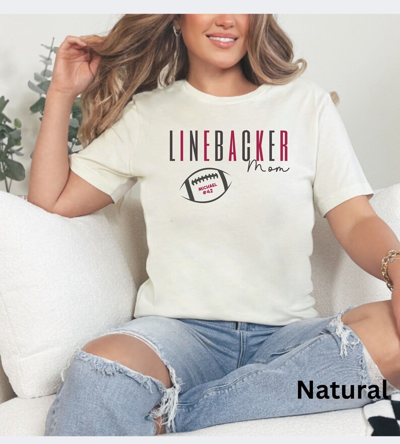 Puede incluir: Una camiseta blanca con el texto "LINEBACKER Mom" y un gr&aacute;fico de f&uacute;tbol en blanco y negro con el texto "MICHAEL #42" en rojo. La camiseta est&aacute; puesta en una persona que lleva pantalones vaqueros azules. La palabra "Natural" est&aacute; escrita en negro en la parte inferior de la imagen.