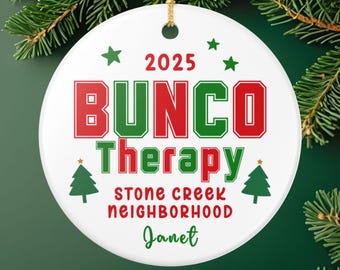 May include: A round white ceramic ornament with a gold hanging string. The text reads 2025 BUNCO Therapy, STONE CREEK NEIGHBORHOOD, and Janet. The design features red and green lettering, three green stars, and two green Christmas tree icons.