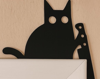 May include: Black cat-shaped metal wall decor holding a knife. The cat has large, round eye holes and is perched above a white door frame. The background is a light tan color.