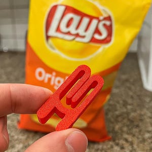 May include: A red chip bag clip is held in front of a yellow and orange Lay's potato chip bag. The clip is shaped like the letter "H" and is designed to seal the bag. The bag has the Lay's logo and the word "Original" printed on it.